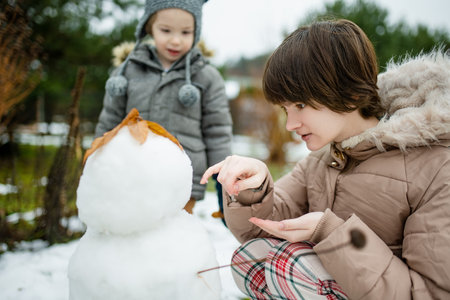 Adorable teenage girl and her little brother building a snowman in the backyard. Cute children playing in a snow. Fun winter activities for family with kids.の写真素材