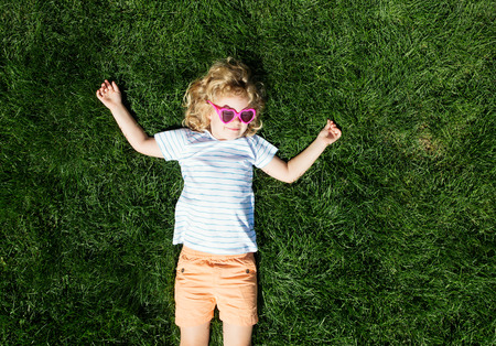Portrait of a smiling little girl in heart shaped sunglasses lying on green grass