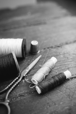 Sewing accessories: bobbins of thread, scissors, needle, thimble on wooden table. Black and white photo. Tailoring and sewing conceptの写真素材