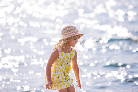 Cute little girl in yellow dress and straw hat standing barefoot on the waves along the beach on a summer sunny dayの写真素材