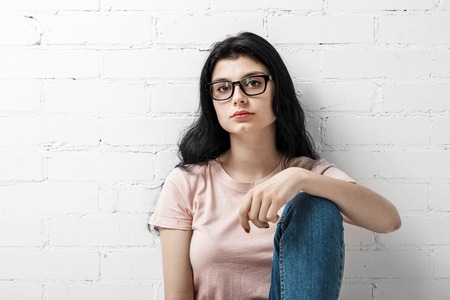Portrait of smart beautiful brunette girl in eyeglasses with natural make-up sitting near white...