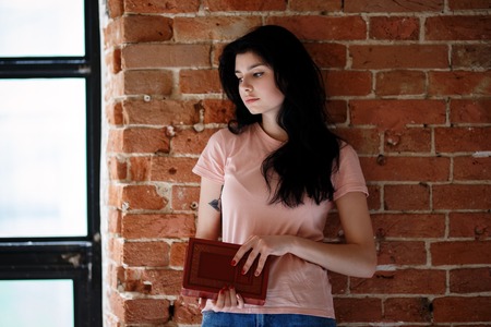 Beautiful brunette young woman with book standing near brick wall.の写真素材