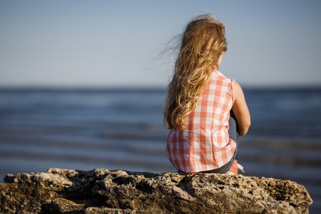 Little girl sits on a rocky shore and looks at the sea.の写真素材