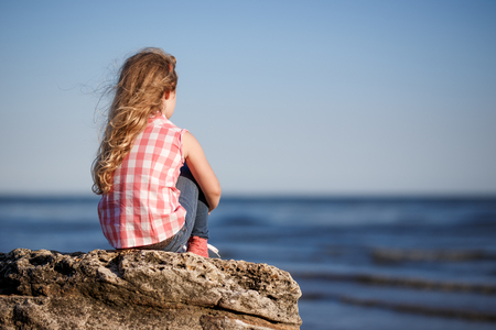 Little girl sits on a rocky shore and looks at the sea.の写真素材