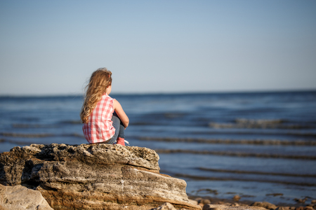 Little girl sits on a rocky shore and looks at the sea.の写真素材