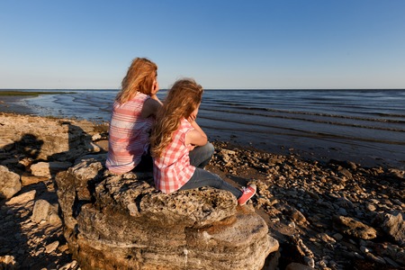 Little girl and her mother sitting on a rocky shore and looking at the sea.の写真素材
