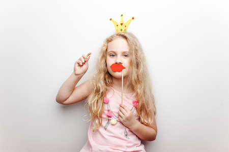 Cute little girl with paper crown and red lips is sitting on red chair at home.の写真素材