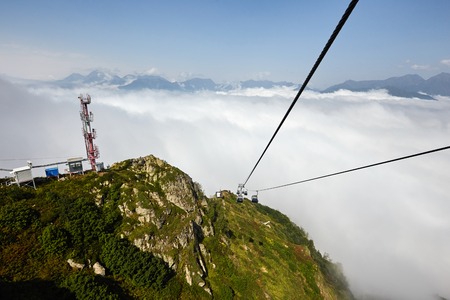 View from top of ropeway with multiple seats and stretched cables above trees in deep fog. Mountains...
