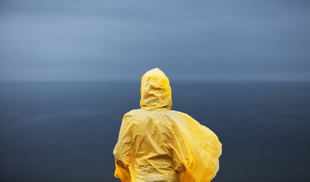 Woman in yellow raincoat looking away on deep blue sea in rainy autumn day.