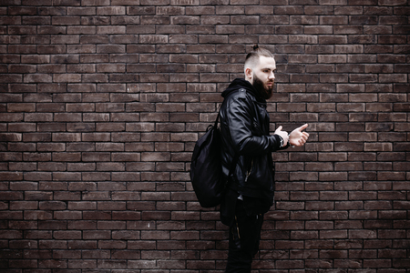 Modern young bearded man in black style clothes posing against brick wall.の写真素材