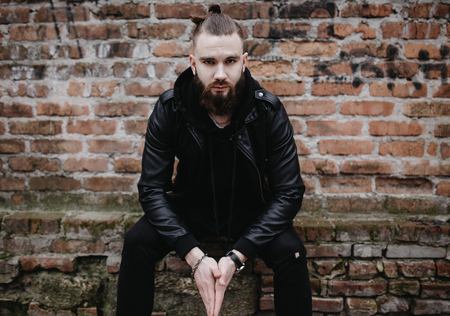 Modern young bearded man in black style clothes posing against brick wall.の写真素材