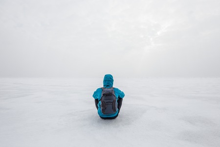 Man with backpack sitting alone on the lake ice and staring forward. Cold cloudy winter day