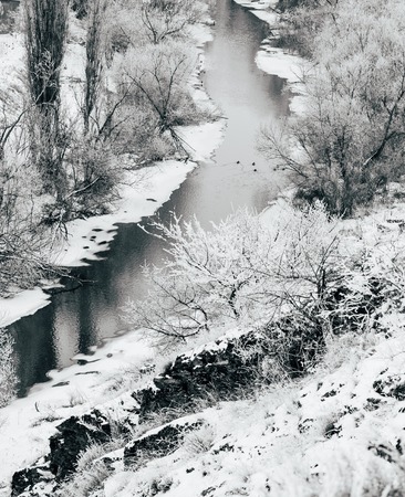 A snow-covered landscape, half frozen river with trees on the banks, flows through a shallow valley....