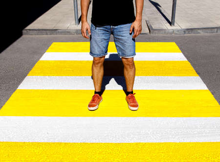 A young man in a black T-shirt and denim shorts stands at a pedestrian crossing.の写真素材