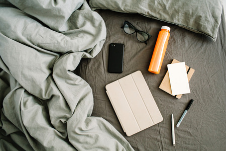 Tablet, diary, mobile, fresh juice, glasses in bed with grey sheet and pillows. Flat lay, top view home office background.の写真素材