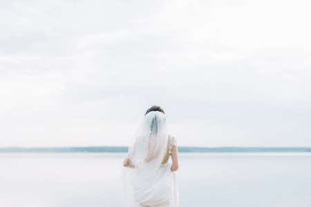Back view on bride in wedding dress look at horizon near lake. Minimal bridal fashion background.の写真素材