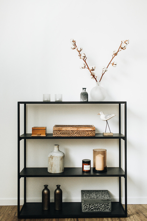 Modern nordic interior design concept. Stylish black rack with caskets, cotton branch, vase, candles and bird figurine at white wall. Interior blog lifestyle composition.の写真素材