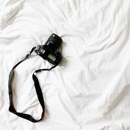 A black camera lying at bed with whit linen. Minimal background. Business , photography concept.