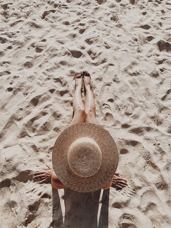 Summer travel fashion and vacation concept. Beautiful young, tanned woman with a straw hat is lying and relaxing on tropical beach with white sand showing legs. Top view, flat lay.の写真素材