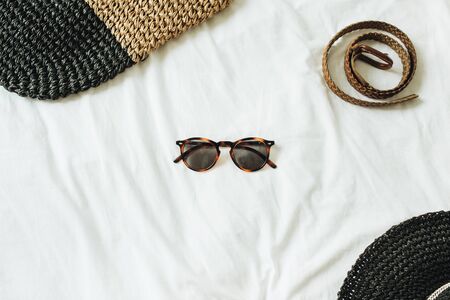 Women's fashion accessories with glasses in the middle of the photo and belt, straw hat, straw bag lying on bed with white sheet. Summer and travel concept. Top view, flat lay.の写真素材