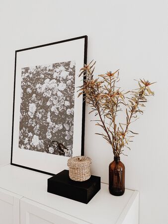 A beautiful painting with black and white flowers and black frame, a little straw basket, black box, dried flowers in brown vase made of glass standing on white chest of drawers. Lifestyle concept and Scandinavian design.の写真素材