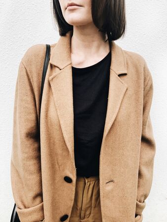 Autumn,fall and fashion concept. A young woman with brown hair, beige coat, black shirt, brown trousers standing against white wall. Closeup photo with neutral colors.の写真素材