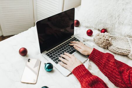 Christmas / New Year composition. Young woman hands in red knitted sweater typing on laptop on the white bed with white blanket decorated with christmas red and blue balls and winter knitted hat. Minimal business freelance concept.の写真素材