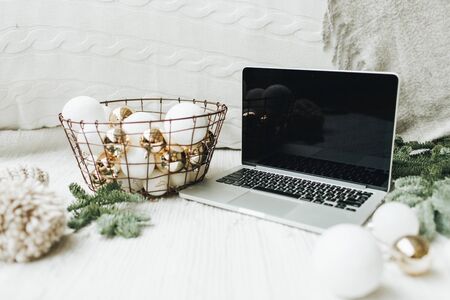 Christmas / New Year / xmas composition. Mock up, copy space. Laptop lying on the white blanket decorated with christmas tree branches, festive basket of white and gold balls, warm hat. Business freelance concept.の写真素材
