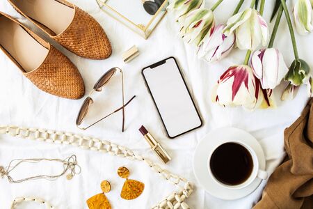 Fashion / beauty blogger home office desk. Blank screen smart phone, tulip flowers bouquet, clothes and accessories on white linen. Flat lay, top view lifestyle composition.の写真素材