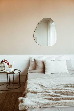 Stylish beige color bedroom with bed, linen, pillows, mirror, bedside table with red berries bouquet, book. Modern classic interior design. Comfortable home living room.の写真素材