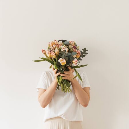 Young pretty woman in white blouse holding tulip flowers bouquet in hands against white wall. Holiday celebration festive floral conceptの写真素材