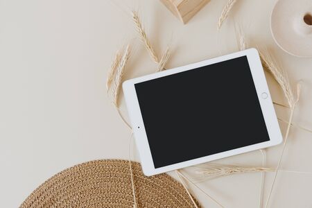 Tablet pad with blank screen on wheat / rye stalks and wicker napkin on beige background. Flat lay,...