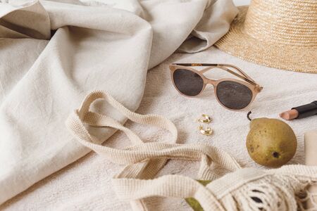 Neutral fashion composition with women's accessories and bijouterie on beige blanket. String bag, straw hat, sunglasses, rings, earrings, pear. Minimal lifestyle concept.の写真素材