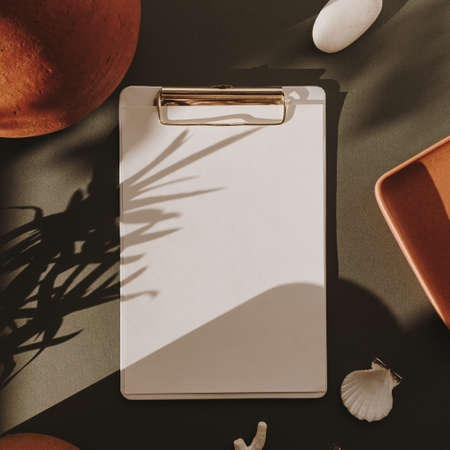 Blank paper sheet clipboard pad with mockup copy space, stones, seashells, coral, candle, clay pot in floral sunlight shadows on deep green background. Flat lay, top view empty templateの写真素材