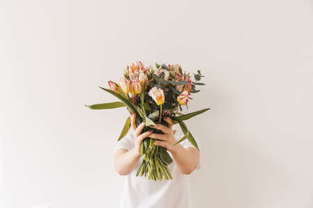 Beautiful girl in hold in hands flower bouquet against white wall. Minimal beauty / fashion festive backgroundの写真素材