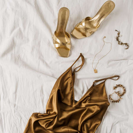 Sexy fashion concept with women's clothes and accessories. Golden shoes, brown silk dress, golden necklace, bracelet, earrings on white linen. Flat lay, top view minimal feminine vogue background.の写真素材