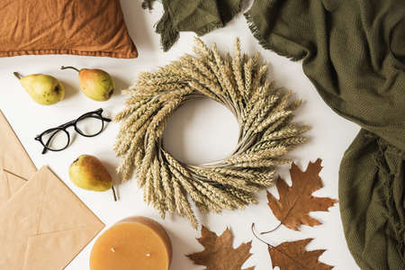 Wreath made of wheat straw lying on white table with blanket, pillow, glasses, pears, envelops, dry...