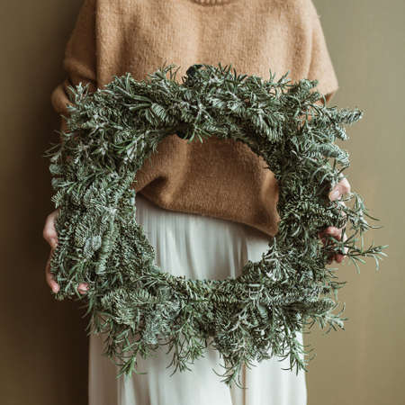 Young beautiful woman holding round wreath frame made of fir branches against olive wall. Minimal fashion festive Christmas / New Year celebration concept.の写真素材
