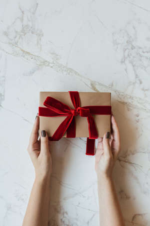 Handmade paper gift box in women hands on marble background. Minimal festive Christmas, New Year celebration concept. Flat lay, top view.の写真素材