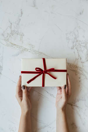 Female hands hold handmade paper gift box on marble background. Minimal festive Valentine's Day / Christmas / New Year celebration concept. Flat lay, top view.の写真素材