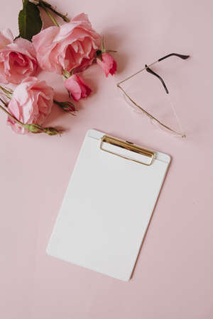Blank paper sheet clipboard note pad, rose flowers bouquet, glasses on pink background. Minimal home...