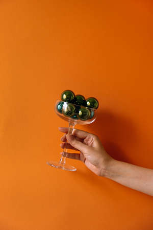 Female hand holding wineglass with green Christmas baubles. Christmas / New Year concept. Minimal...