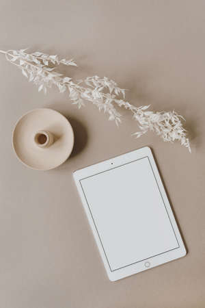 Blank screen tablet, white floral branch on neutral pastel beige background. Flat lay, top view...