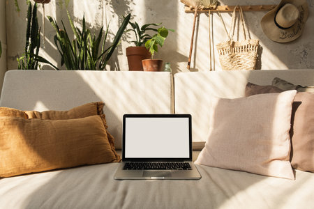Laptop with blank screen on comfortable sofa in warm sunlight shadows. Minimal boho styled interior...