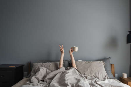 Woman laying in bed, holding mug with coffee in hand and show "Victory" symbol. Happy morning breakfast concept. Freelancer, pandemic quarantine day lifestyle concept.の写真素材