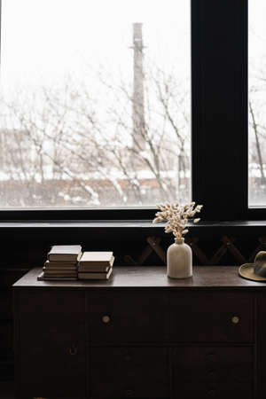 Rabbit bunny tail grass bouquet, books stack on solid wooden table against window. Aesthetic dark home interior design concept. Beautiful floral foliage composition.の写真素材