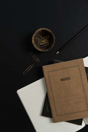 Flatlay of notebooks, clips in wooden bowl on black background. Home office desk workspace....