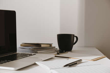 Laptop on table with coffee cup, paper sheet against white wall. Minimalist home office workspace.の写真素材