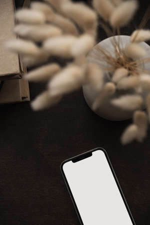 Aesthetic flatlay of blank screen mobile smart phone, books stack, bunny rabbit tail grass bouquet on deep brown background. Blog, social media, web, magazine copy space template. Flat lay, top view.の写真素材