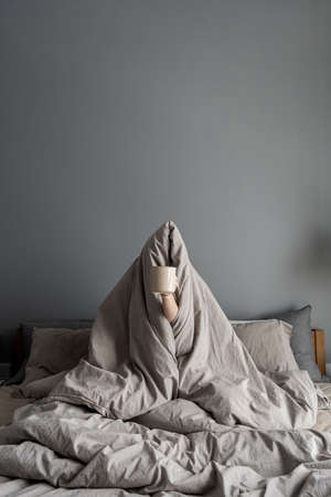 Woman sitting in bed, holding mug with coffee in hand. Happy morning breakfast concept. Freelancer, pandemic quarantine day lifestyle concept. Minimalist bed room interior design.nの写真素材